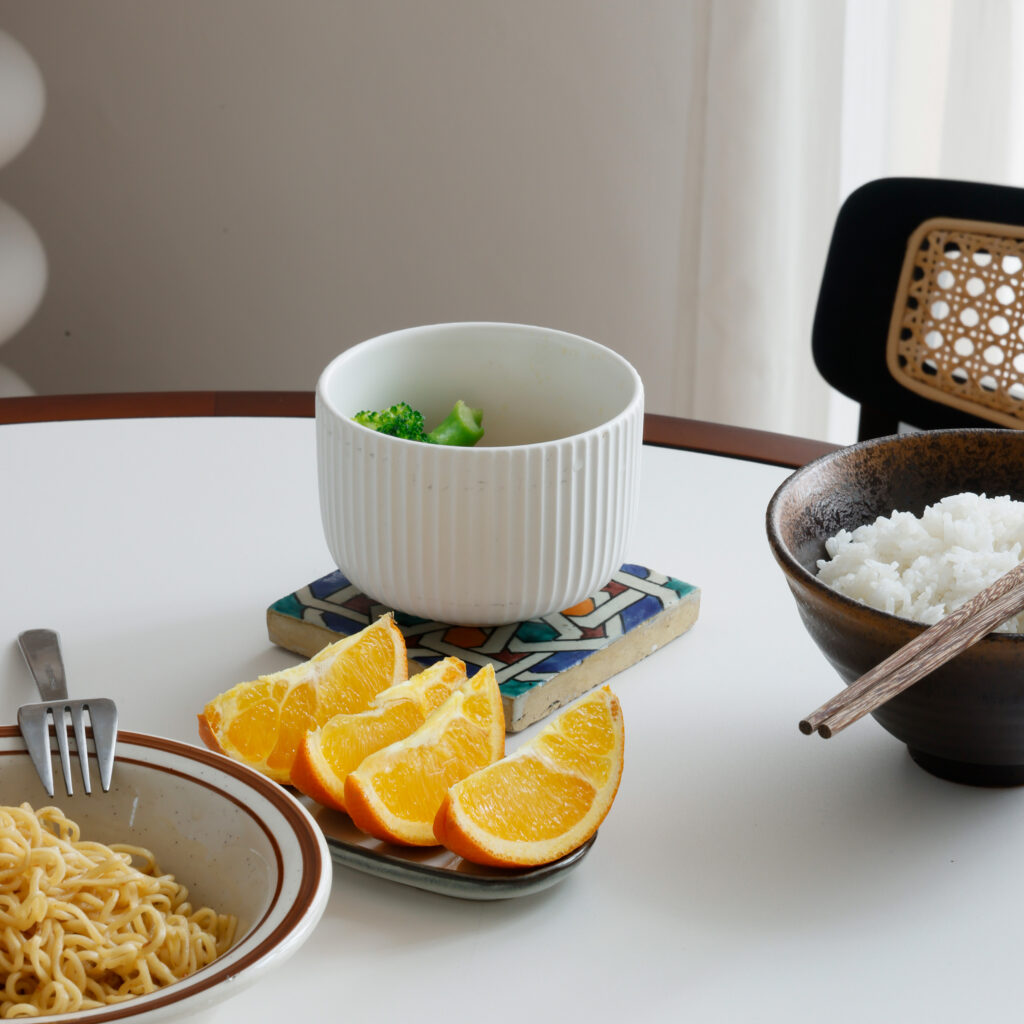 A table set with a bowl of white rice and chopsticks, a plate of orange segments, a bowl of soup with broccoli, and a plate of ramen noodles with a fork.