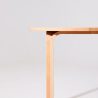 A close-up view of a modern wooden table with a rounded edge and a single leg, set against a plain light grey background.