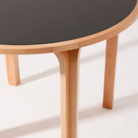 A close-up of a round table with light wooden legs and a smooth, black tabletop, set against a white background.