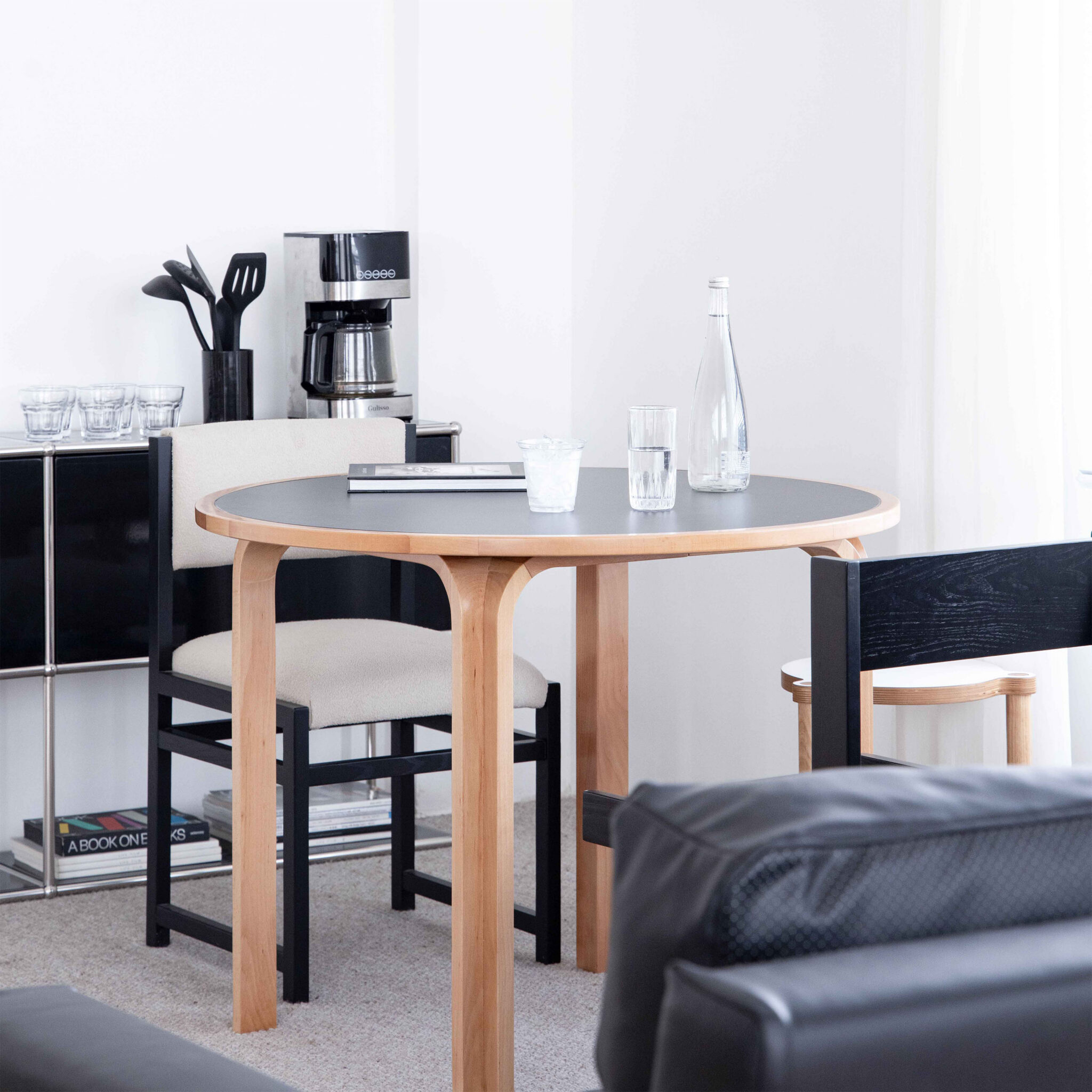 A modern dining area with a round wooden table, two chairs, a glass bottle, glasses, and a coffee maker on a shelf in a bright, minimalist room.