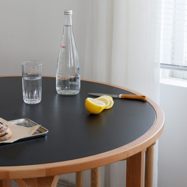 A round wooden table with a black top holds a glass of water, a clear bottle, a knife, sliced lemon, and a tray with pastries. A window with white blinds and sheer curtains is in the background.