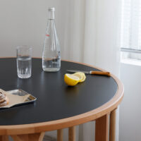 A round wooden table with a black top holds a glass of water, a clear bottle, a knife, sliced lemon, and a tray with pastries. A window with white blinds and sheer curtains is in the background.