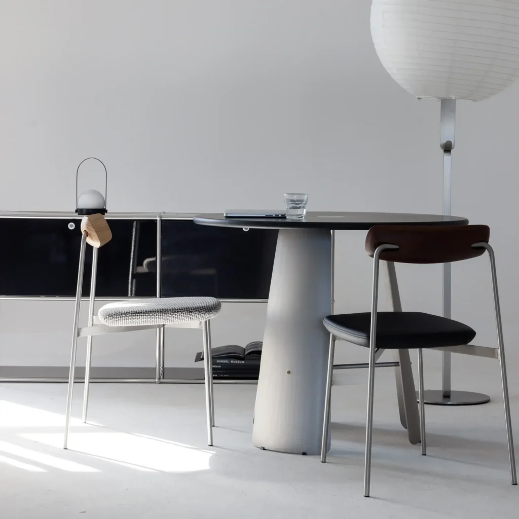 A minimalist room with a round table, two modern chairs, a glass of water, a floor lamp with a paper shade, and a sleek cupboard against a plain white wall.
