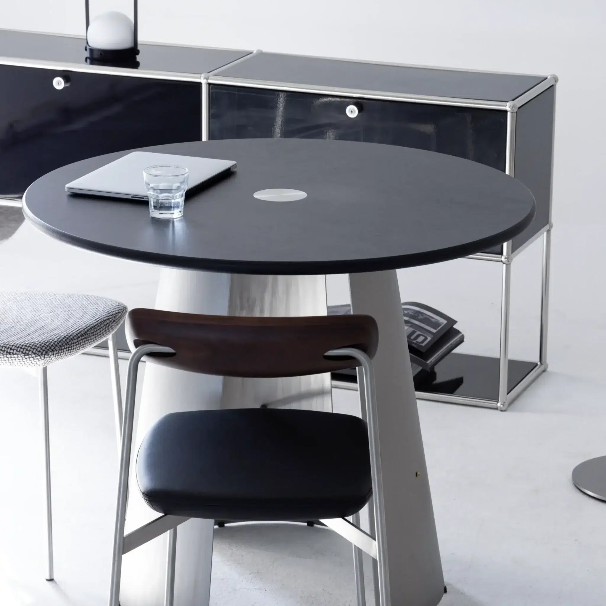 A modern minimalist office set-up with a round black table, a black and wood chair, a closed laptop, and a glass of water. In the background, there is a sleek black sideboard with chrome edges and a black rotary telephone.