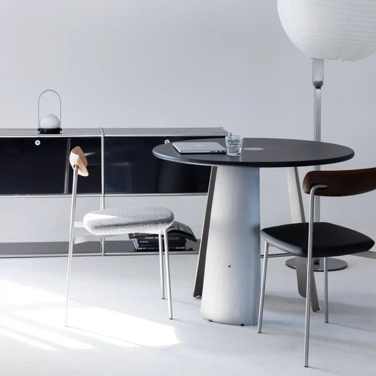 Monet Round Dining Table_Black_Front View(14) A modern minimalist room with a round black table, two chairs, a glass of water, a laptop, a sideboard, and a floor lamp with a white paper shade. The space is bright with a clean, neutral colour palette.