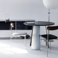 A modern minimalist room with a round black table, two chairs, a glass of water, a laptop, a sideboard, and a floor lamp with a white paper shade. The space is bright with a clean, neutral colour palette.
