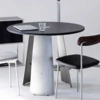 A round black table with a silver base holds a closed laptop and a glass of water. Two modern chairs, one with a dark seat and back, and one with a grey patterned cushion, are nearby in a minimalistic setting.