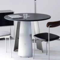 A round black table with a silver base holds a closed laptop and a glass of water. Two modern chairs, one with a dark seat and back, and one with a grey patterned cushion, are nearby in a minimalistic setting.