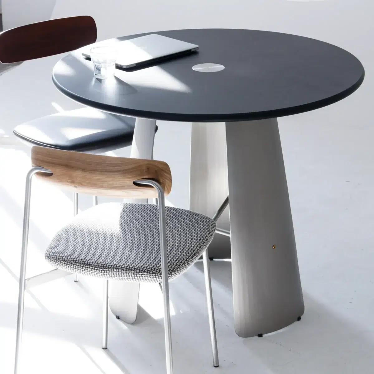 Monet Round Dining Table_Black_Front View(5) A round black table with a glass of water, a closed laptop, and two modern chairs, one with a wooden backrest and the other with a dark backrest, in a bright, minimalist room.