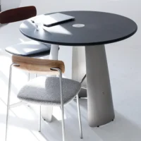 A round black table with a glass of water, a closed laptop, and two modern chairs, one with a wooden backrest and the other with a dark backrest, in a bright, minimalist room.