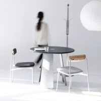 A minimalist room with a round black table, two modern chairs, a coat stand, and a large paper lantern. A person in motion walks past, blurred against the white background. A glass sits on the table.