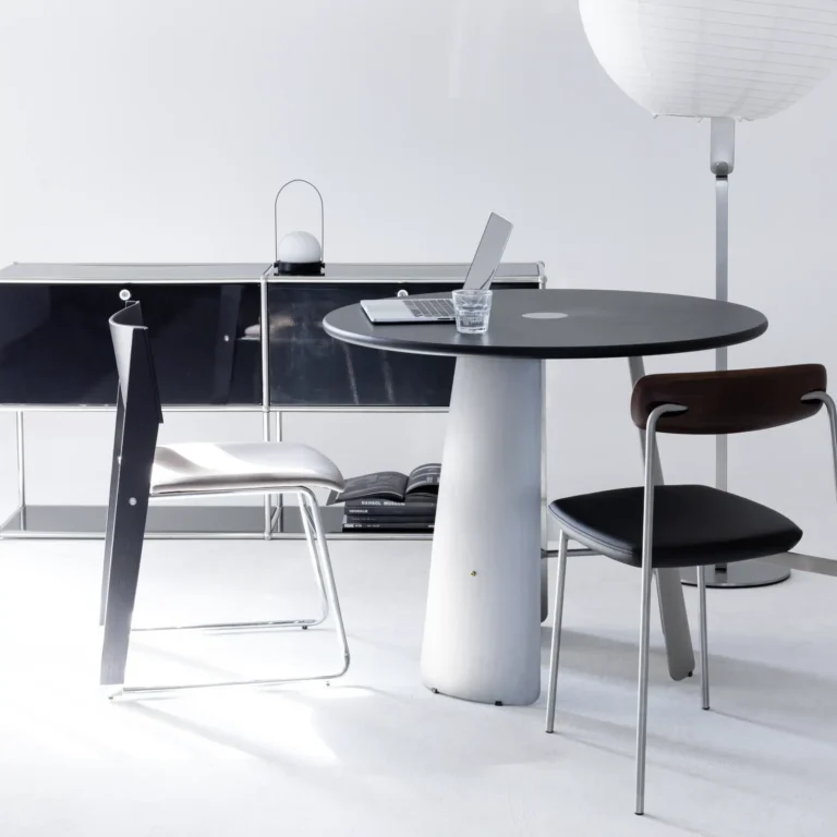 A modern minimalist office set-up with a round black table, two chairs, a closed laptop, a glass of water, a black sideboard, and a tall white floor lamp in a bright, white space.