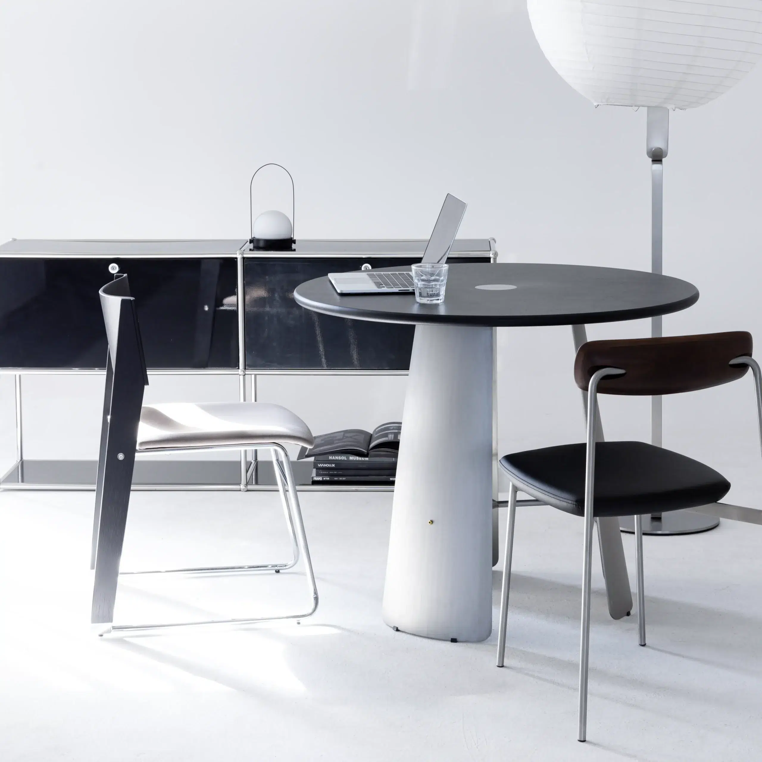 A modern minimalist office set-up with a round black table, two chairs, a closed laptop, a glass of water, a black sideboard, and a tall white floor lamp in a bright, white space.