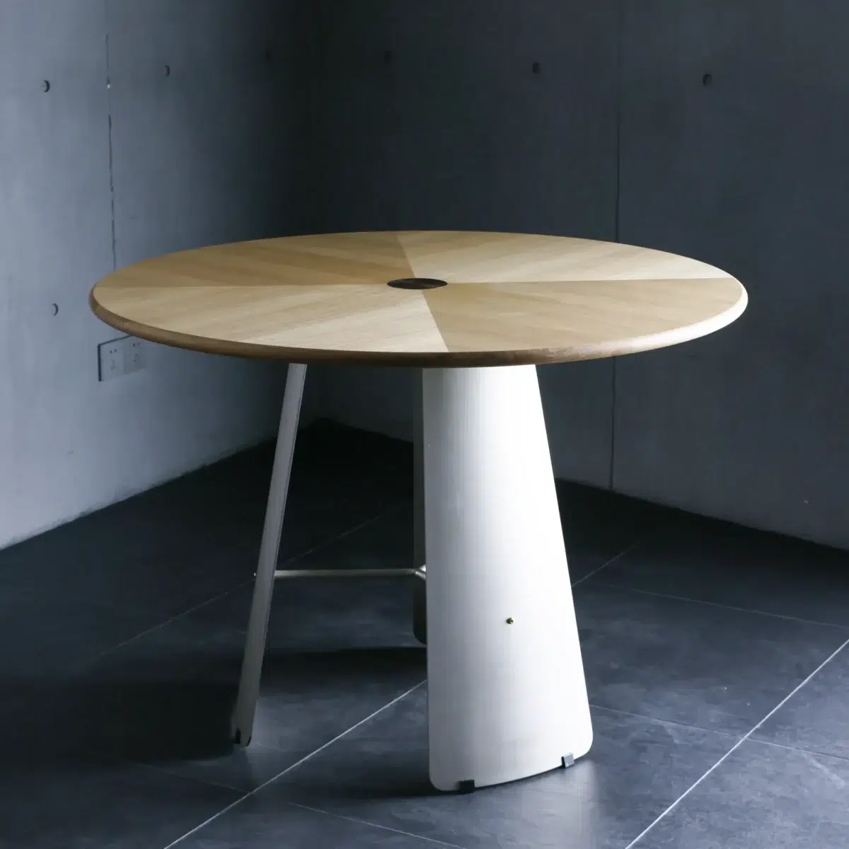 Monet Round Dining Table_Front View A modern round wooden table with a light-coloured top and a single sturdy, white, cone-shaped base stands on a dark tiled floor against a grey concrete wall.