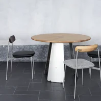 A round wooden table with a white base stands on a dark tiled floor, accompanied by two modern chairs with metal legs—one with a black seat and back, the other with a grey textured seat and light wood backrest.