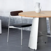 A modern round table with a light wooden top and metal base stands on a dark tiled floor next to a minimalist black chair. A white sculptural vase with handles sits on the table. The background wall is textured grey.