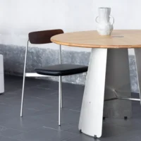 A modern round table with a light wooden top and metal base stands on a dark tiled floor next to a minimalist black chair. A white sculptural vase with handles sits on the table. The background wall is textured grey.