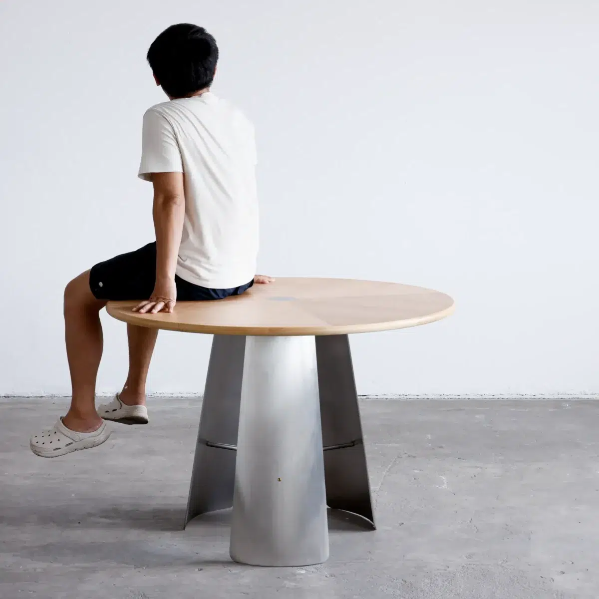Monet Round Dining Table_Front View(5) A person in a white shirt, black shorts, and white clogs sits on the edge of a round wooden table with a metal base, facing away from the camera in a minimalist room.