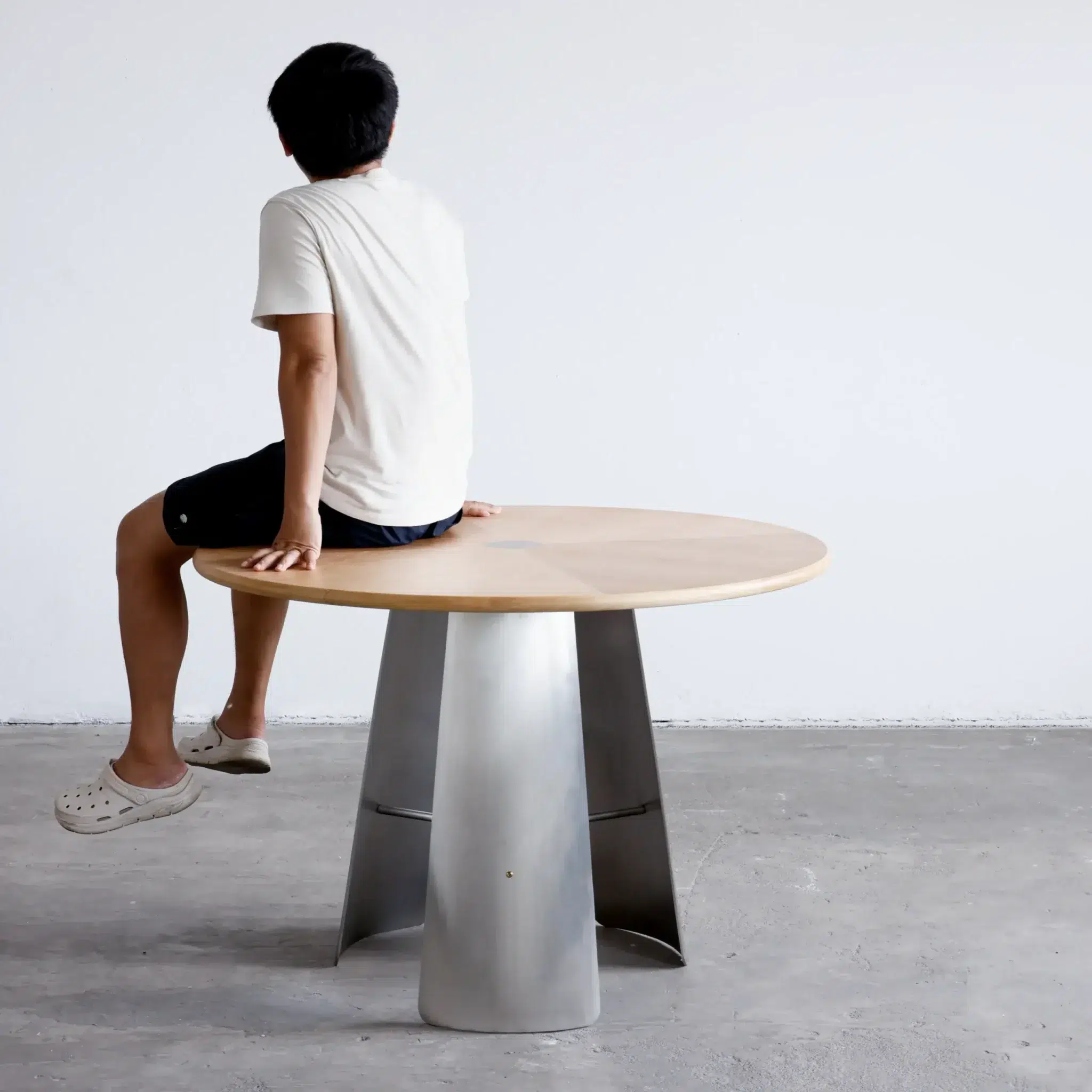 A person in a white shirt, black shorts, and white clogs sits on the edge of a round wooden table with a metal base, facing away from the camera in a minimalist room.