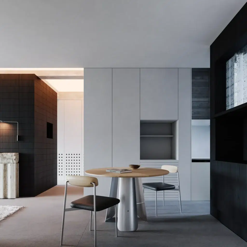 A modern, minimalist dining area with a round wooden table, three chairs, grey walls, built-in cupboards, and dark accents. The space is clean, with simple decor and neutral colours.