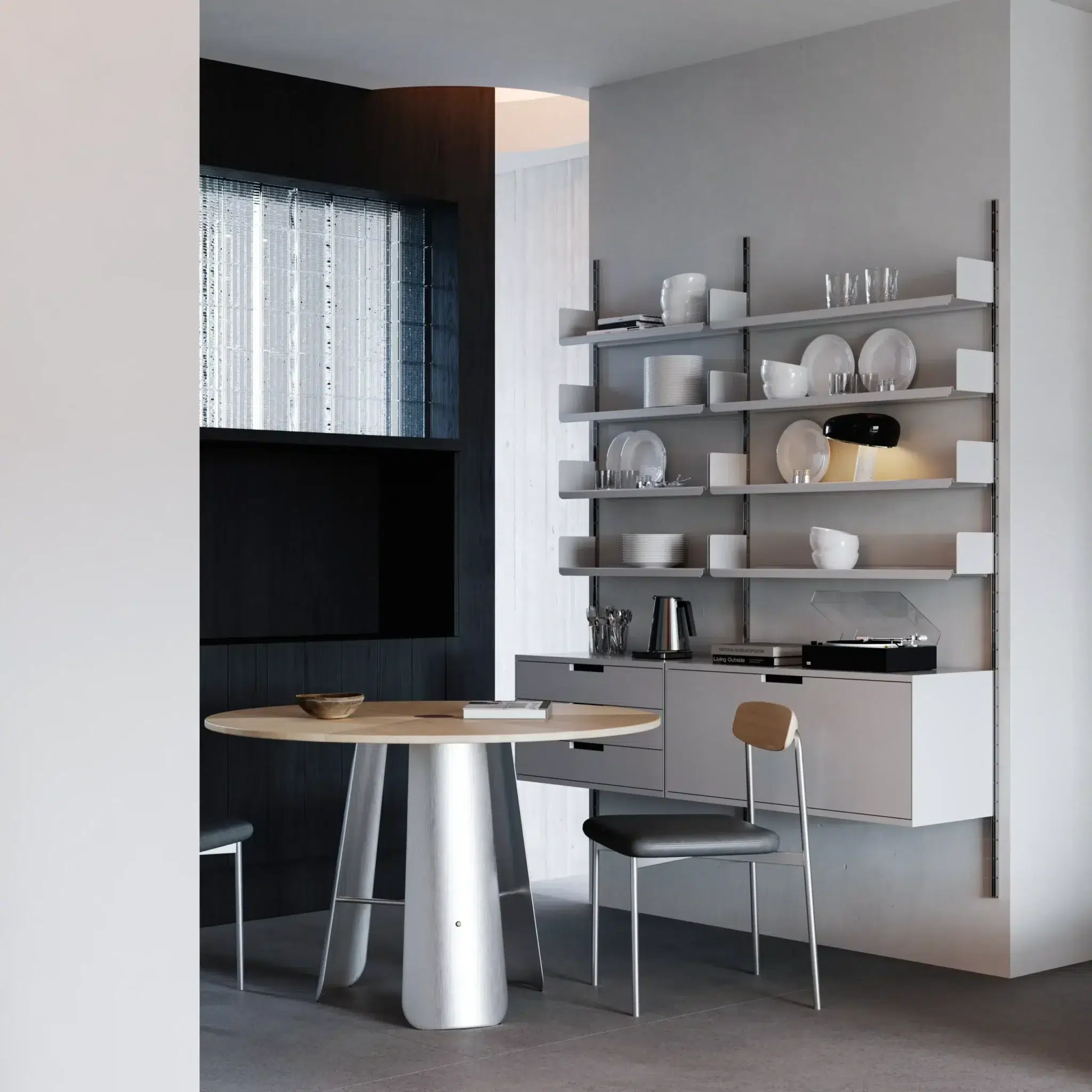 A modern dining area with a round wooden table, two chairs, and wall-mounted shelves holding crockery, glasses, and kitchenware. The space features minimalist design and neutral colours.