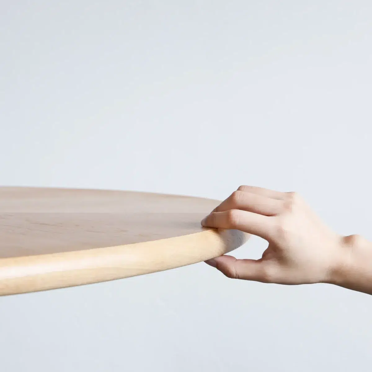Monet Round Dining Table_Front View_Detail A hand gently grasps the edge of a round, light wooden table against a plain, light grey background.