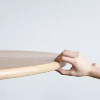 A hand gently grasps the edge of a round, light wooden table against a plain, light grey background.