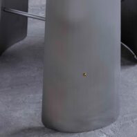 Close-up of a smooth, curved, grey metallic surface with a small, round, gold-coloured dot near the centre, set on a textured concrete floor. Parts of similar metallic structures are visible in the background.