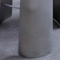 Close-up of a smooth, curved, grey metallic surface with a small, round, gold-coloured dot near the centre, set on a textured concrete floor. Parts of similar metallic structures are visible in the background.