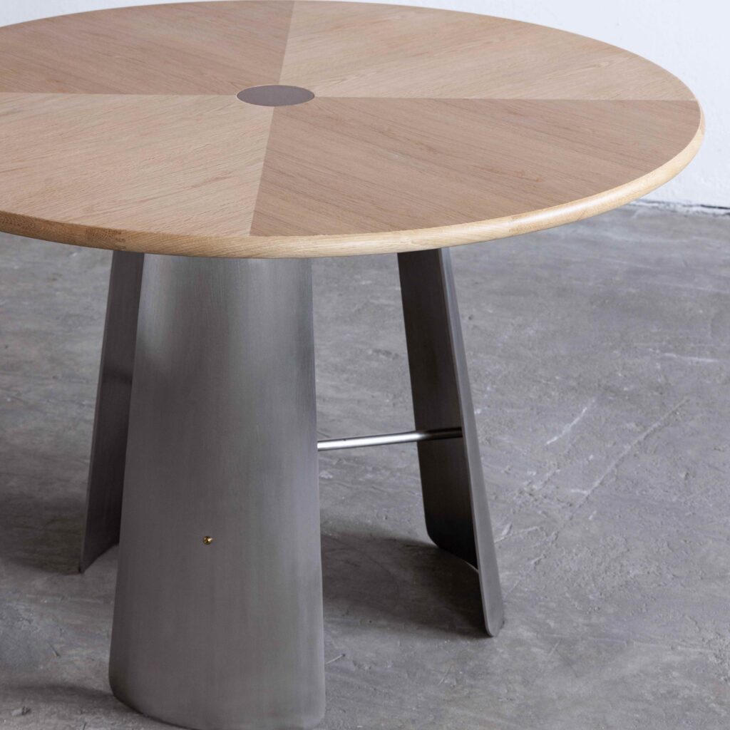 A round table with a wooden top featuring a subtle geometric pattern and a metal base with wide, angled legs, set on a concrete floor.
