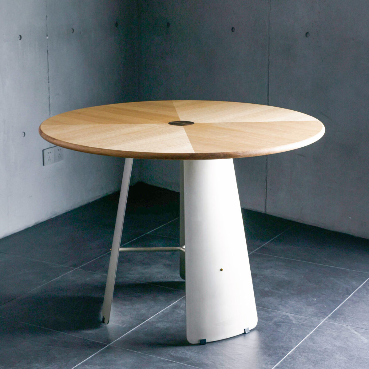 A round wooden table with a geometric pattern on top and a single, wide, white pedestal base, placed on dark tiled flooring against a grey concrete wall.