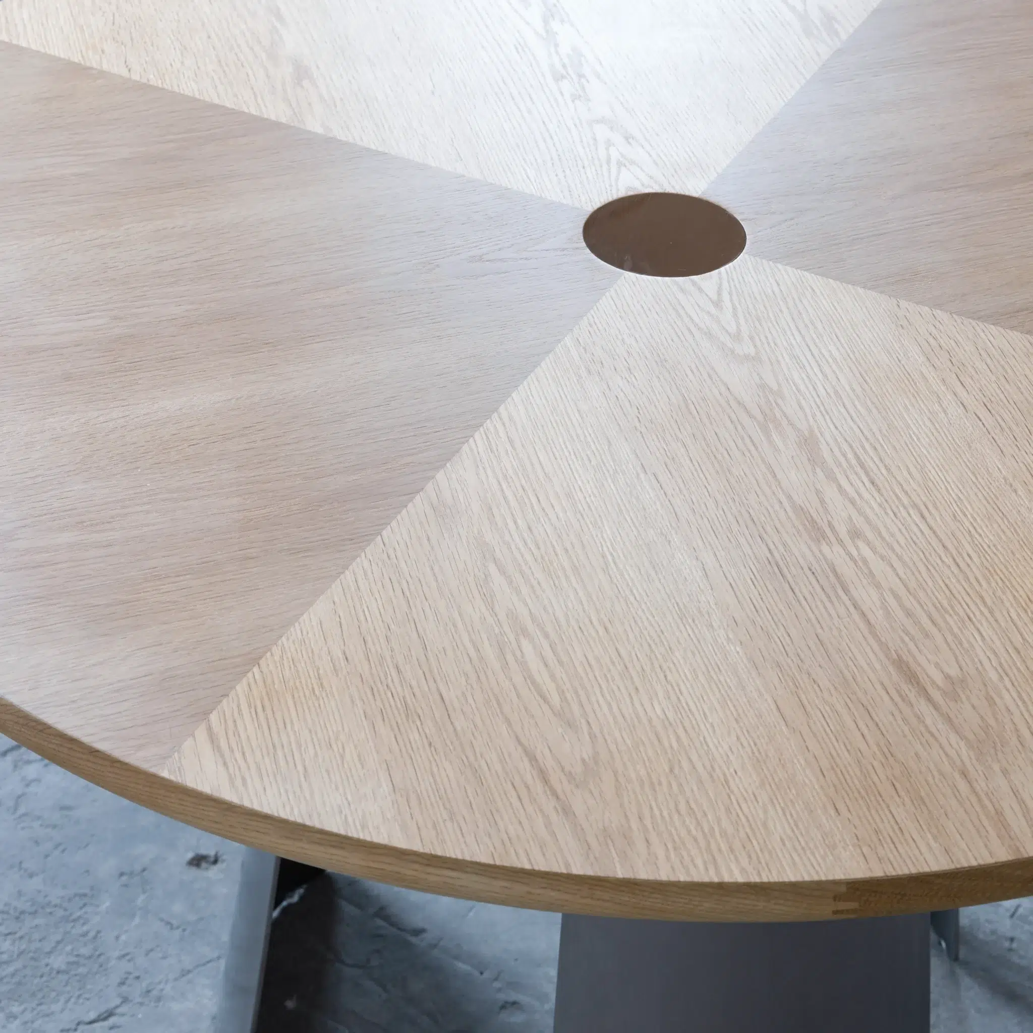 A round wooden table with a geometric pattern of light and medium wood sections and a small circular hole in the centre, standing on a grey base.