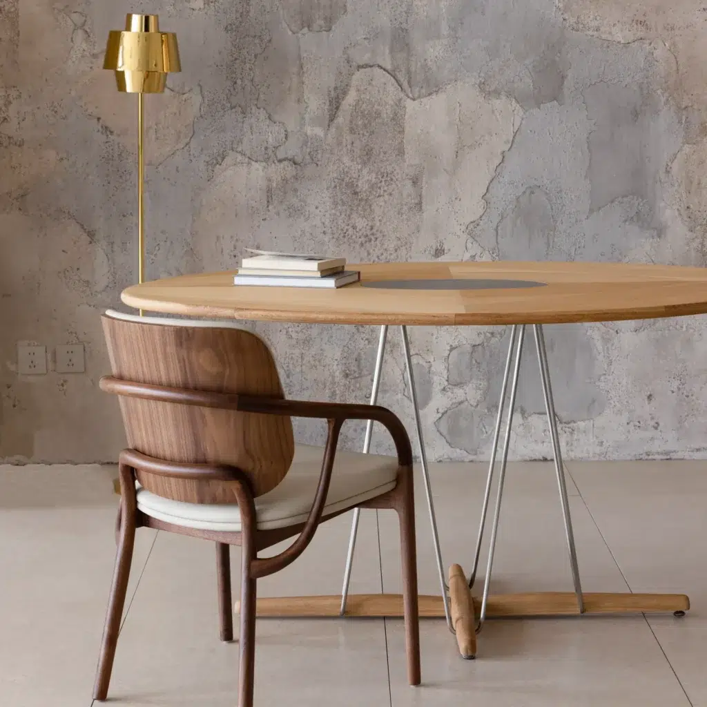 A modern wooden table with metal legs, a stack of books on top, and a cushioned wooden chair sits beside it. A gold lamp stands in the background against a textured, concrete wall.