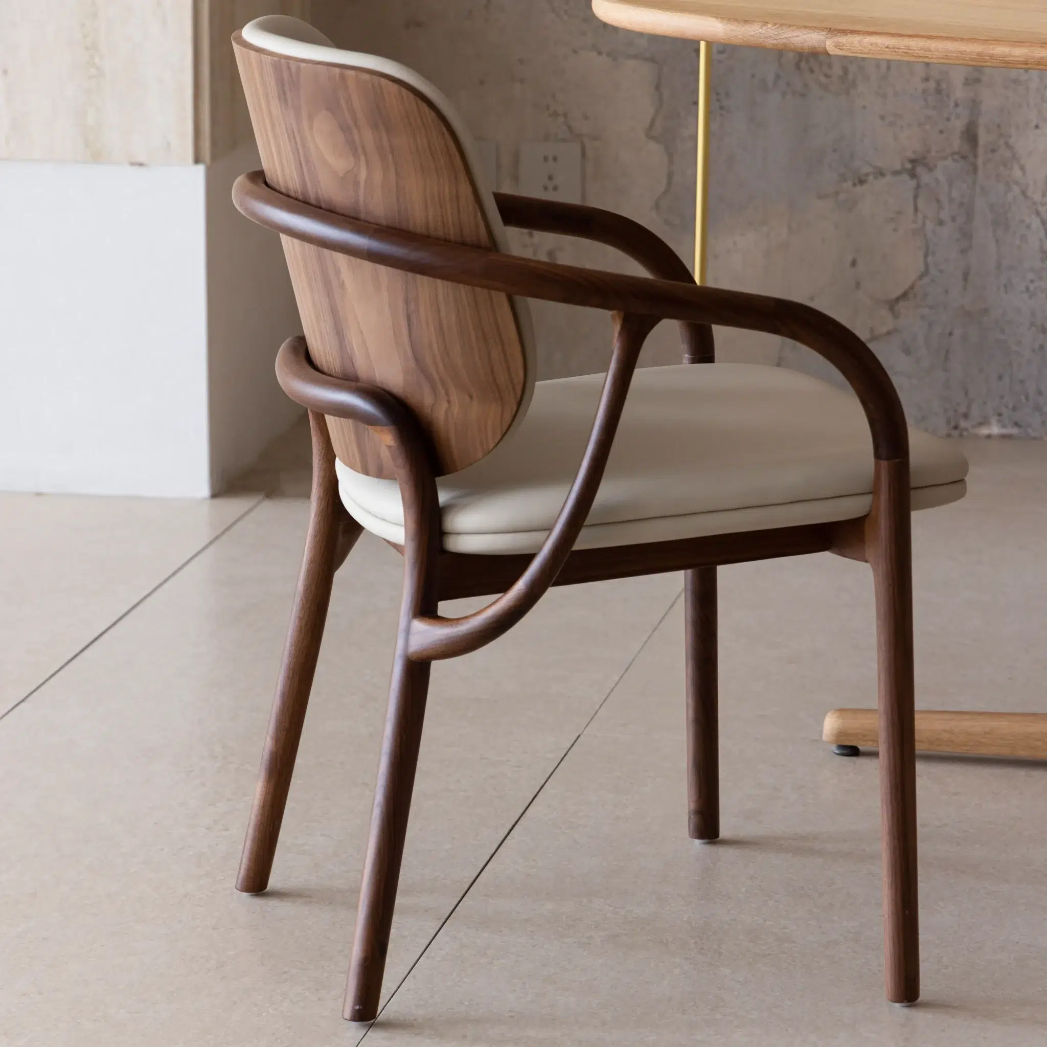 A modern wooden chair with curved arms and legs, featuring a light cushioned seat and backrest, positioned next to a wooden table in a minimal, neutral-toned room.