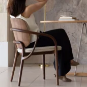 A woman sits sideways on a modern wooden chair with a curved backrest, facing a table with stacked books. She wears a white sleeveless top, black trousers, and beige shoes. The background features a textured wall.