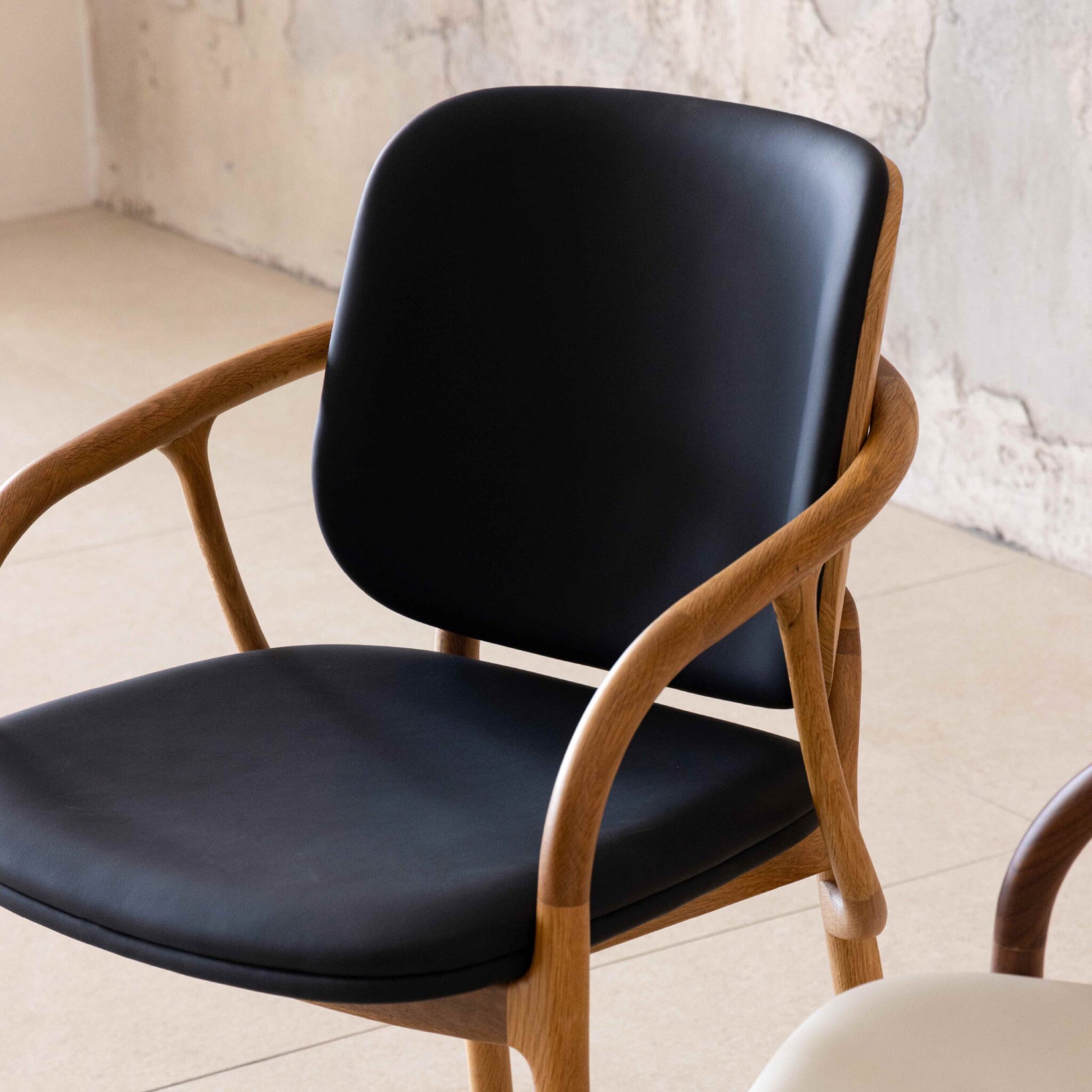 A modern chair with a curved wooden frame and black cushioned seat and backrest, placed indoors near a textured, light-coloured wall.