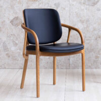 A wooden chair with curved armrests and legs, featuring a black upholstered seat and backrest, is placed on a light-coloured tiled floor against a stone-patterned wall.