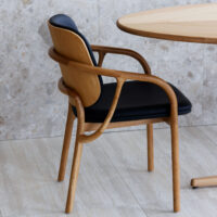 A wooden chair with curved armrests and legs, featuring a black cushioned seat and backrest, is positioned next to a round wooden table against a stone-tiled wall and floor.
