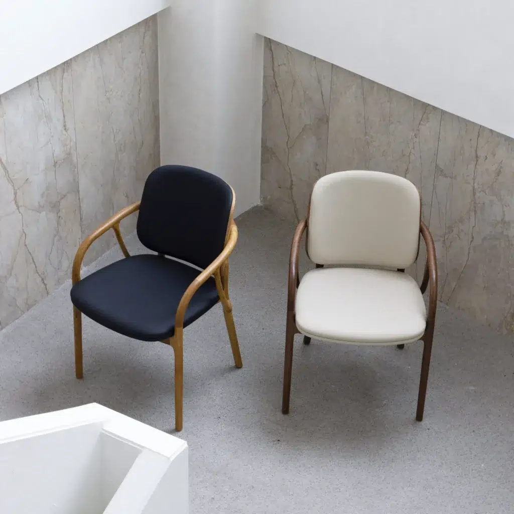 Two modern chairs sit side by side in a minimalist room. One chair has a light wooden frame with black cushions, and the other has a dark wooden frame with cream cushions. Both are on a light-coloured floor near marble walls.