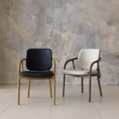 Two modern chairs with wooden frames are placed side by side; one has a black seat and backrest, the other is white. They are set against a textured, light grey wall and light-coloured floor tiles.
