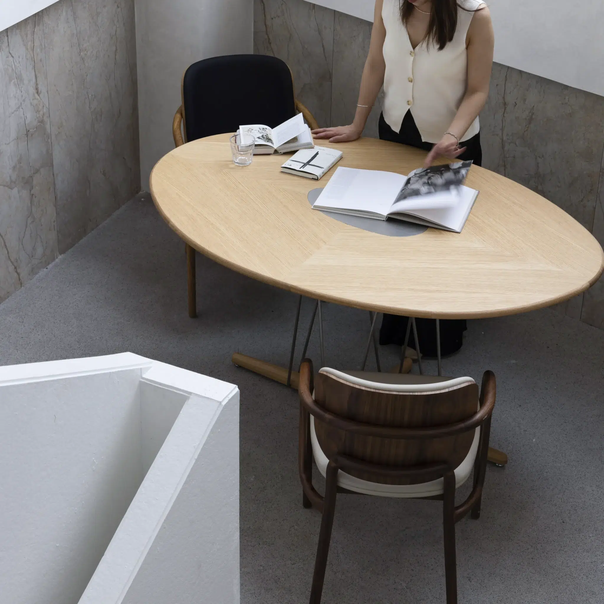 A woman stands at an oval wooden table with open books and a glass of water. Two chairs are positioned at the table, in a modern, minimalist room with light walls and carpeted floor.