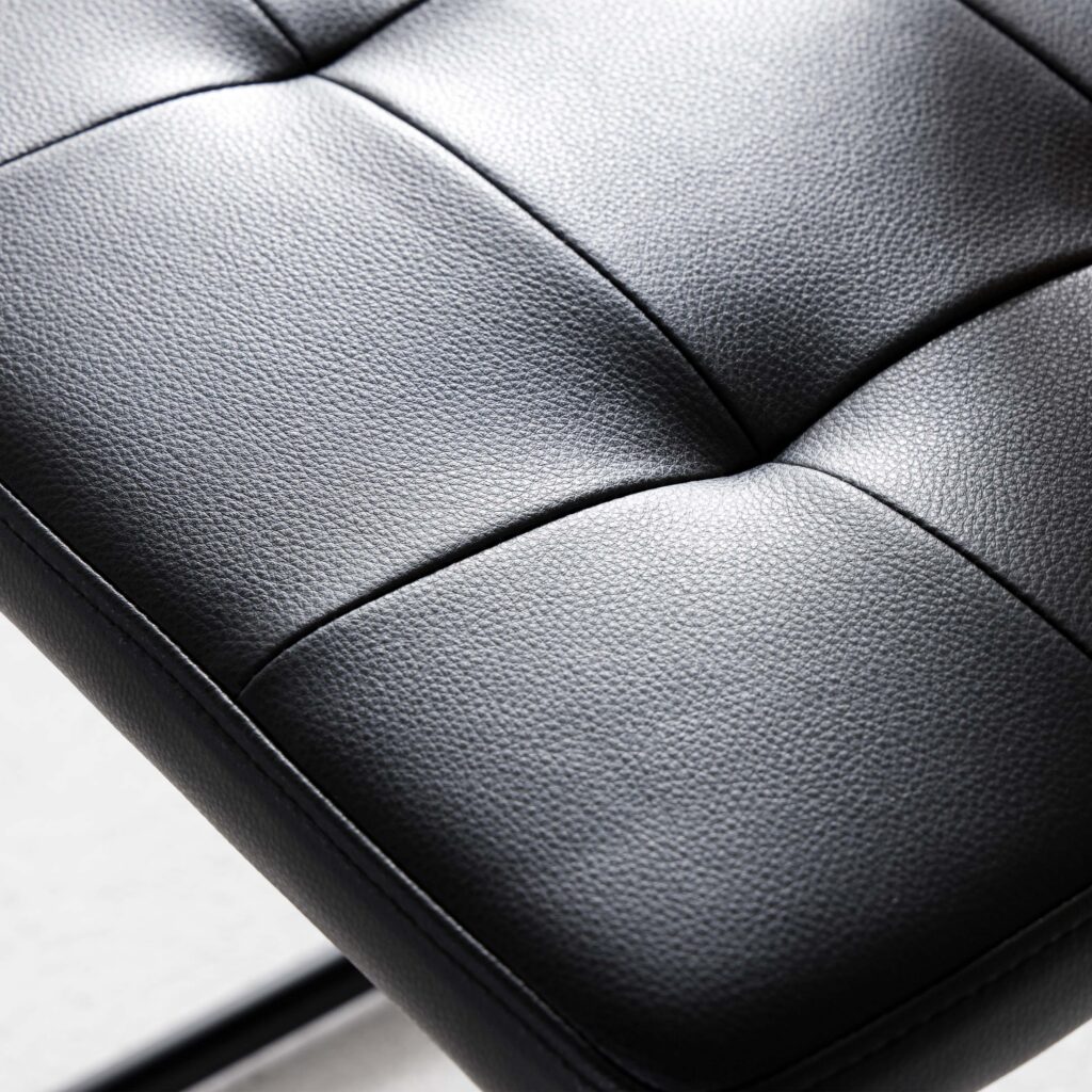 Close-up of a black leather cushion with visible stitching in a square pattern and a textured surface, part of a modern chair or seat.