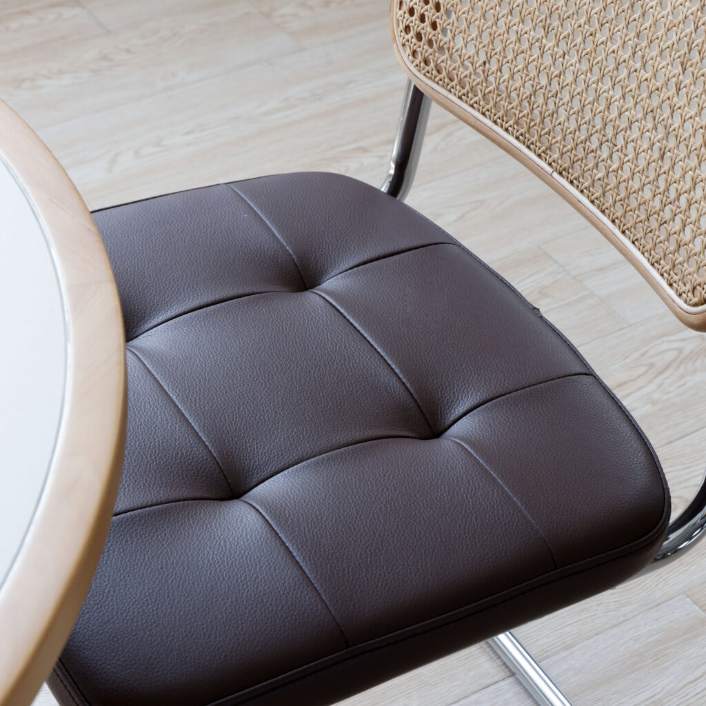 A close-up of a chair with a brown, tufted leather seat and a woven cane backrest, next to a round table with a light wooden edge, set on a light wood floor.