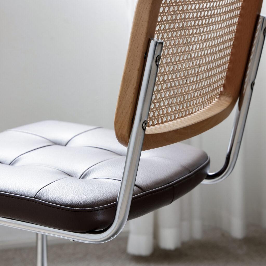A close-up of a modern chair with a tufted, cushioned leather seat, chrome legs, and a wooden backrest featuring a woven rattan design. Light filters through a curtain in the background.