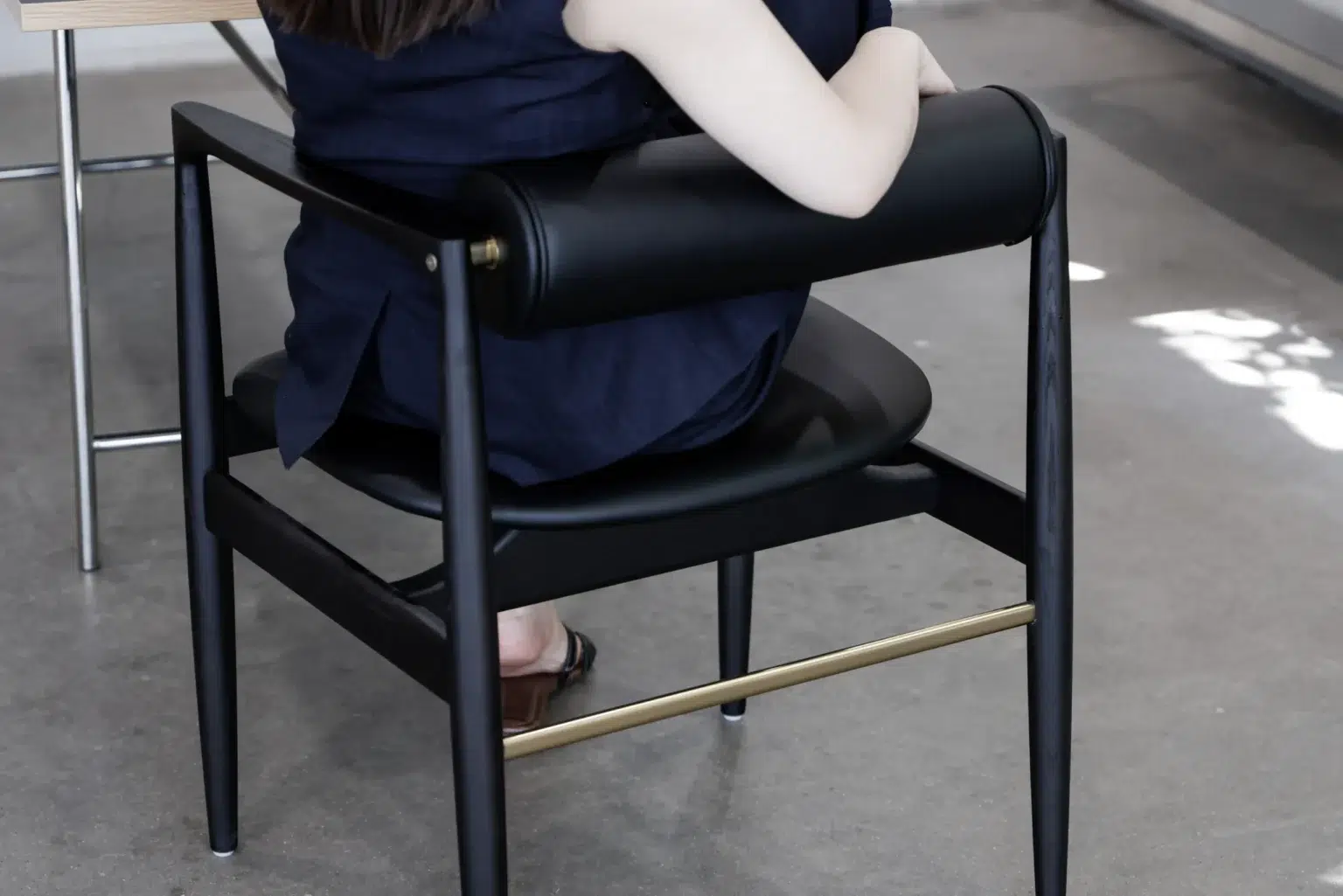 Outdoor02 A person with long dark hair, wearing a navy outfit, sits on a modern black and gold chair at a light wooden table in a room with a concrete floor.