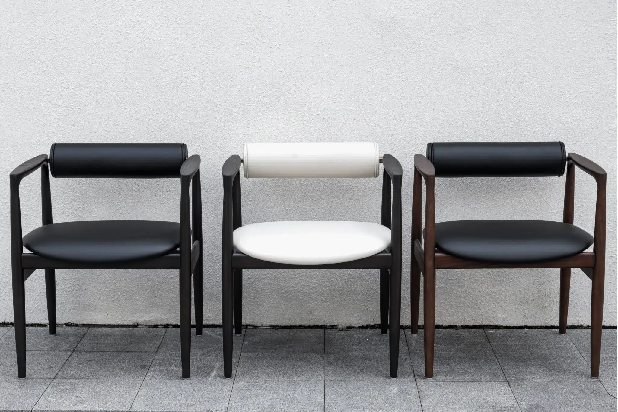 Three modern chairs with wooden frames are placed side by side against a white textured wall. The two outer chairs have black seats and backs, while the middle chair has a white seat and backrest.
