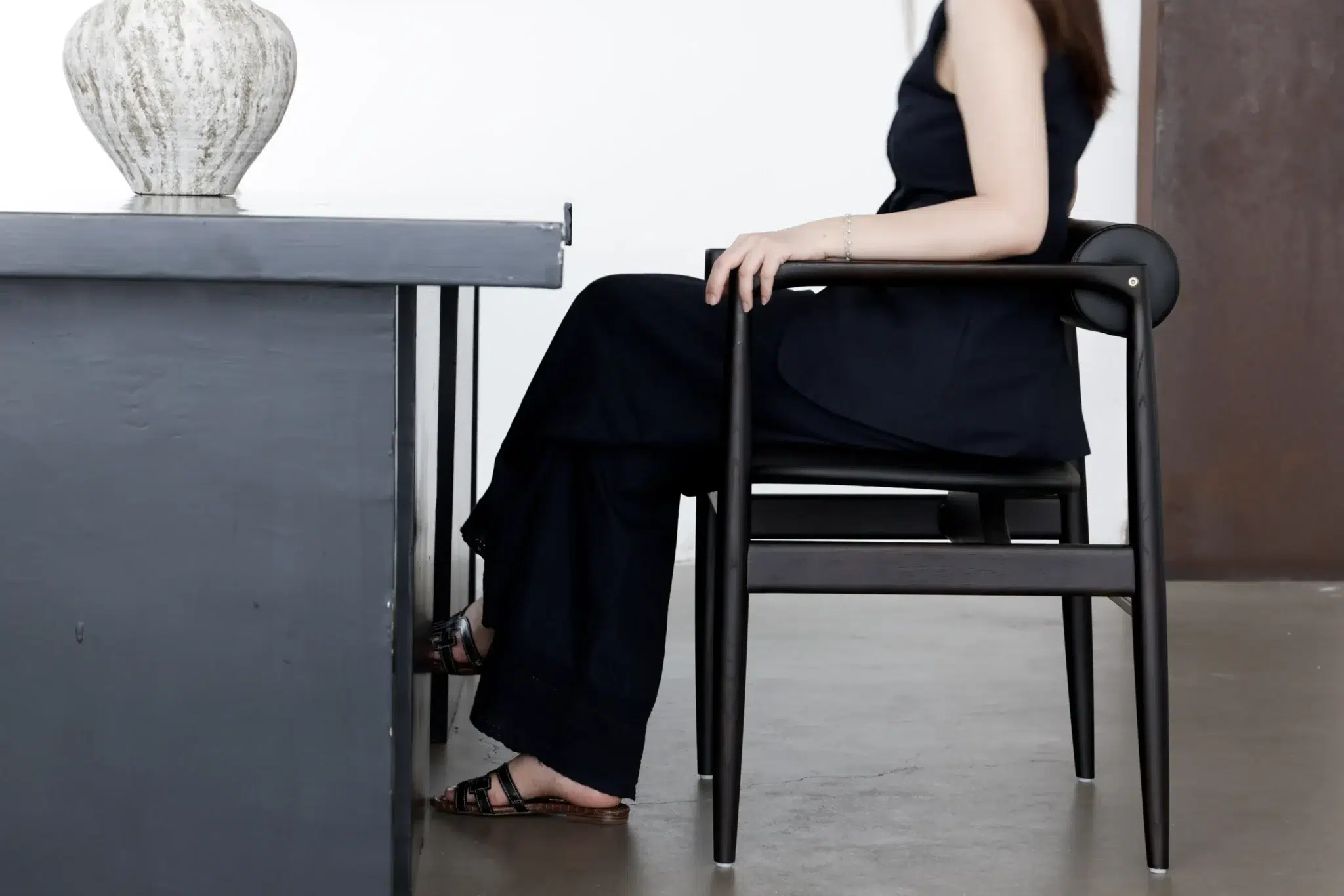 A woman in a black outfit sits sideways on a black chair at a grey desk, with her face out of frame and feet resting on the floor. A light-coloured vase is on the desk. The background is minimal and white.