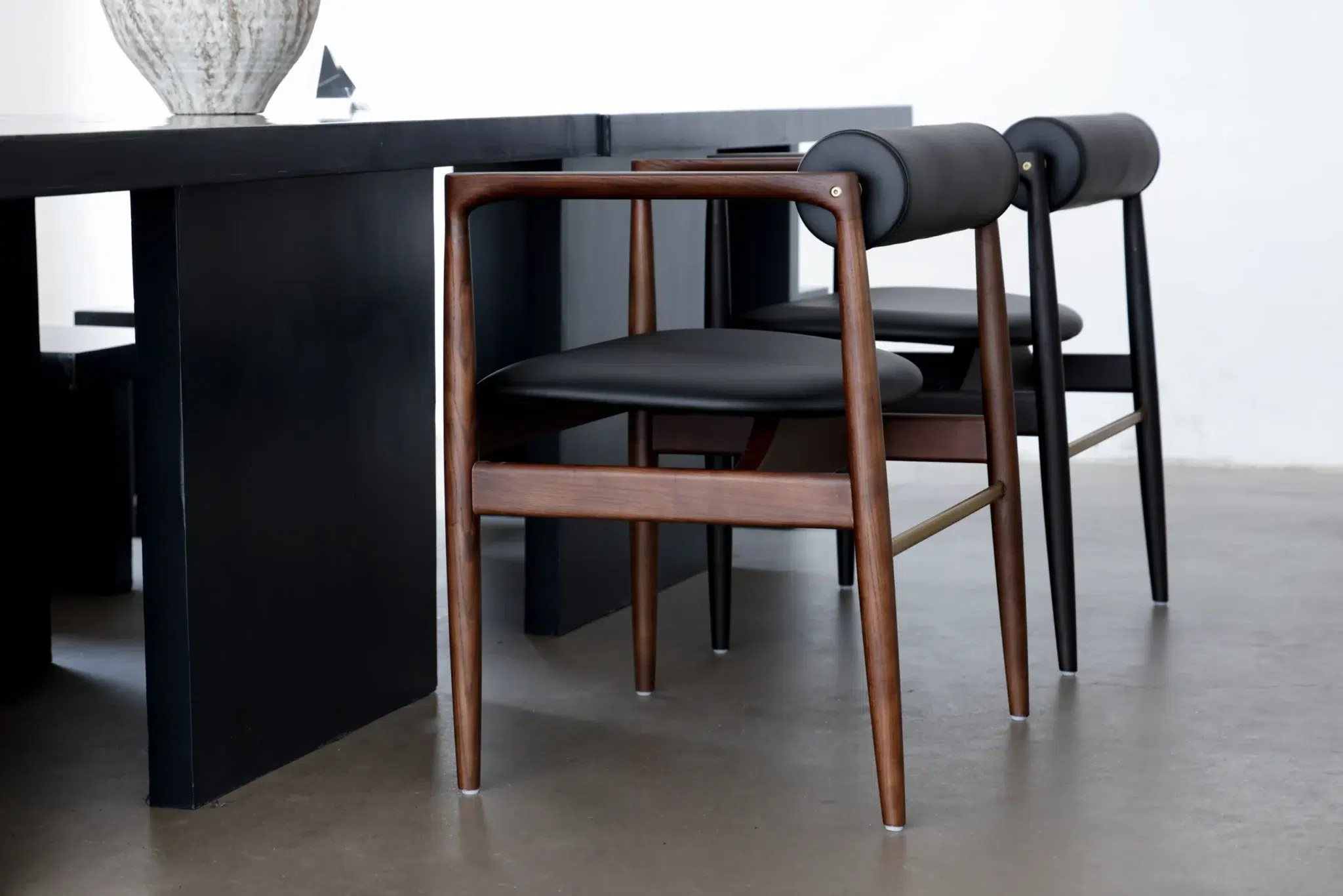Two modern wooden chairs with black cushioned seats and cylindrical backrests are placed next to a sleek black table on a polished concrete floor.