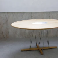 A round wooden table with a white circular centre sits on a unique metal and wood base against a wall with beige marble panels.