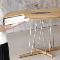 A person holding books stands near a round wooden table with thin metal legs and a dark circular inlay, set against a textured, concrete wall.
